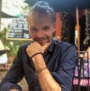 Mężczyzna z brodą siedzi przy stoliku z białym kubkiem, uśmiechając się i opierając podbródek na dłoni. Ma na sobie granatową koszulę i bransoletkę, a sznurkowe lampki i kredowe menu sugerują zbliżające się KONCERTY lub WARSZTATY w przytulnym otoczeniu.