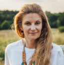 Kobieta o długich, jasnobrązowych włosach ma na sobie białą koszulę i kolorowy naszyjnik z koralików z motywem tygrysa. Katarzyna Poręba stoi na zewnątrz przy dużym kamieniu, zieleni i drzewach - idealna na KONCERTY lub WARSZTATY na łonie natury.