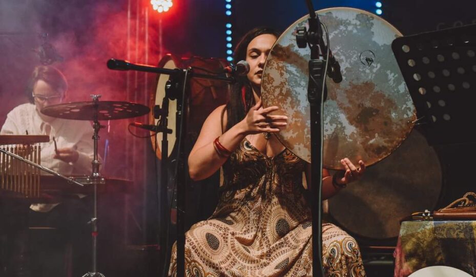 Katarzyna Poręba we wzorzystej sukience siedzi na scenie, grając na dużym bębnie i śpiewając do mikrofonu. W tle muzyk gra na perkusji, otoczony kolorowymi światłami KONCERTY i efektami dymnymi.