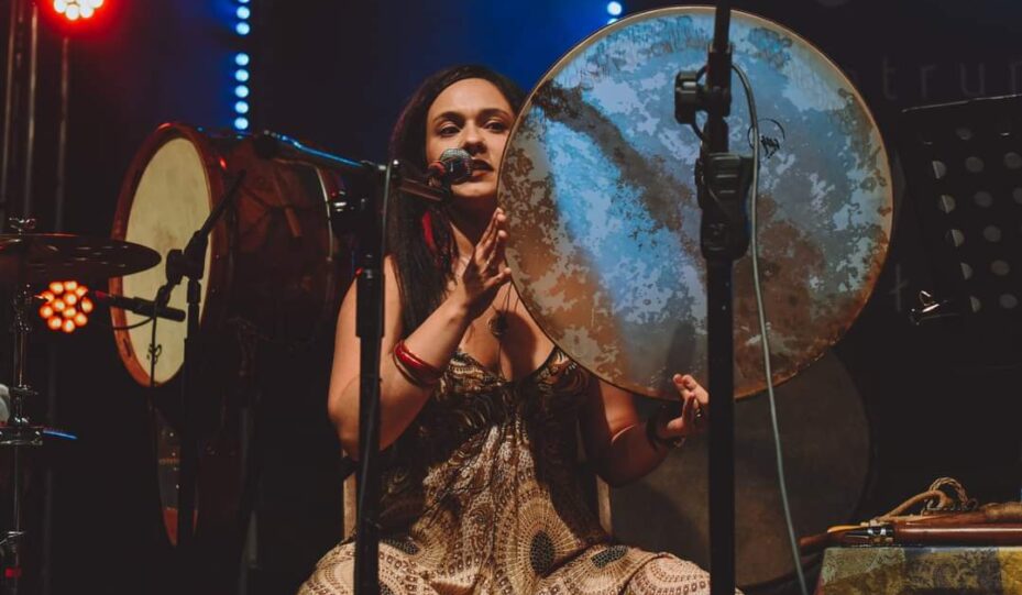 Katarzyna Poręba siedzi na scenie we wzorzystej sukience, gra na dużym bębnie i śpiewa do mikrofonu. Kolorowe światła i widoczna perkusja tworzą scenę dla jej niezapomnianego doświadczenia KONCERTY.